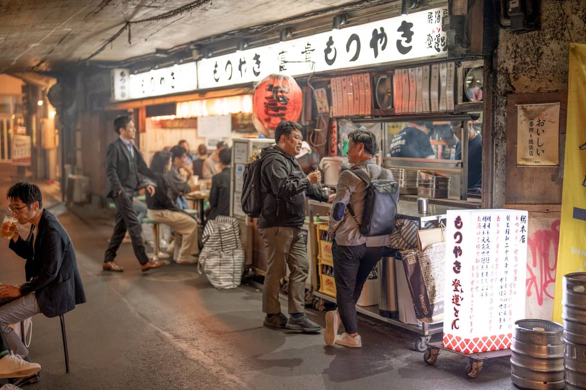 Men standing and chatting at the counter of a traditional Tokyo izakaya under a railway track at night, surrounded by locals eating and drinking.