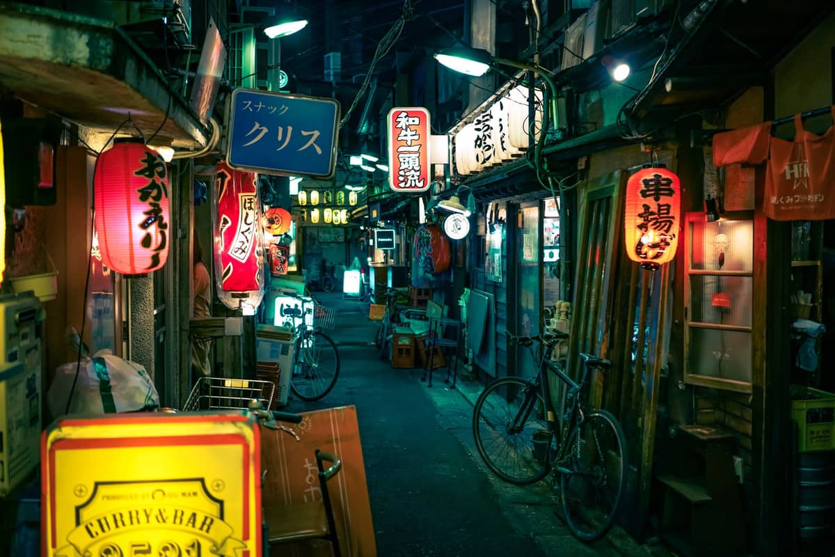 Narrow Tokyo alley at night lined with glowing lanterns, izakayas, bars and bicycles, evoking authentic late-night nightlife atmosphere.