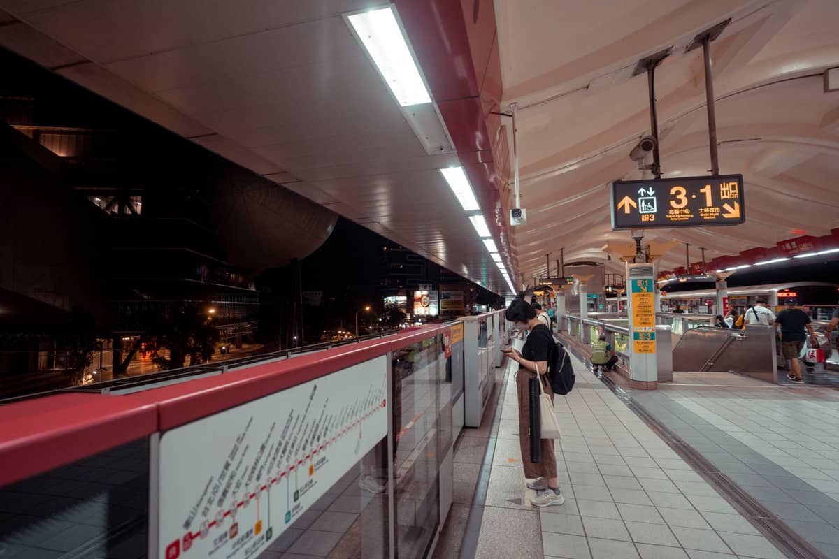 Nighttime scene at a Tokyo train station platform with a timetable sign, person waiting, and city outside.