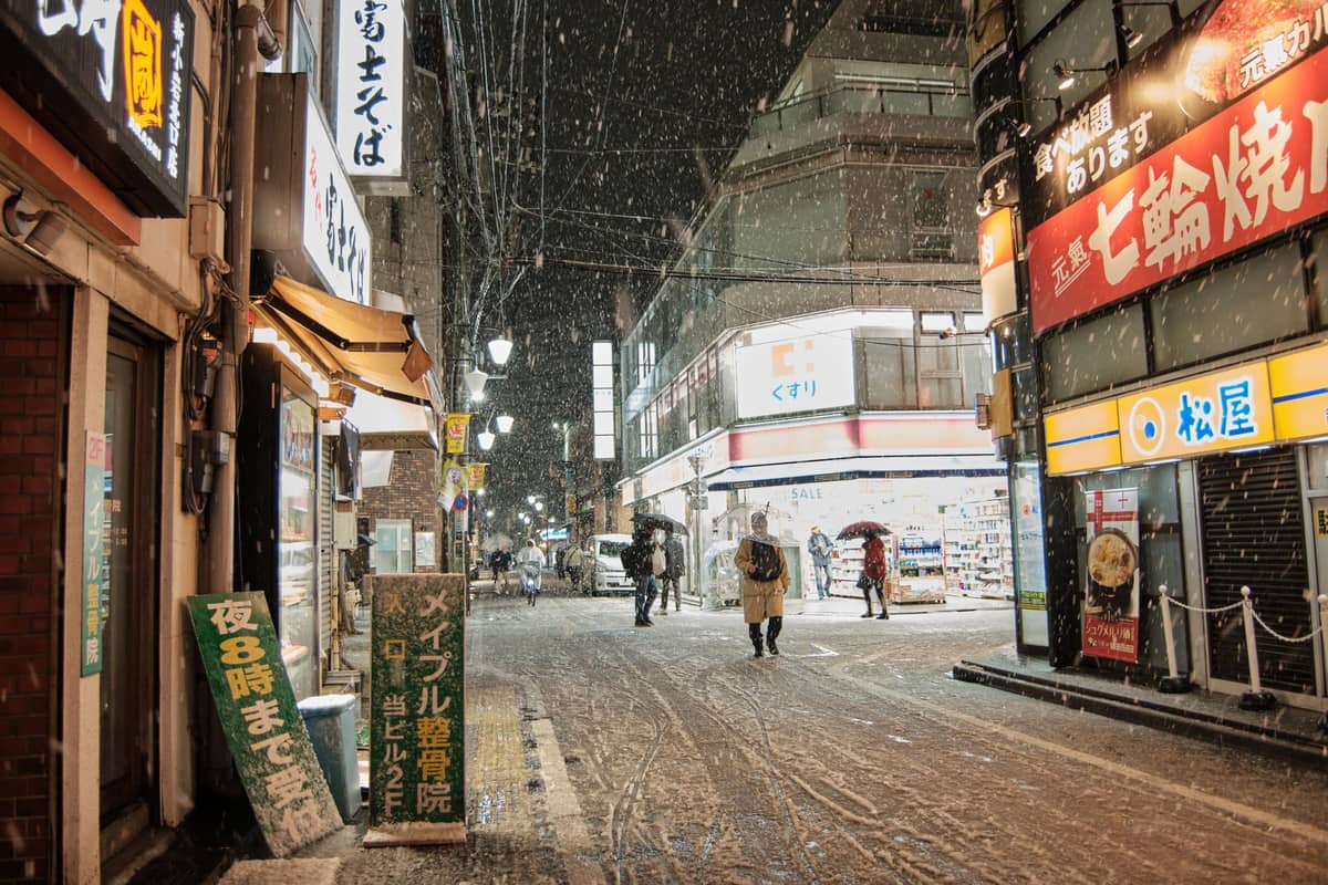 Tokyo street scene at night with people walking, snow falling, and store lights illuminating the wintry setting