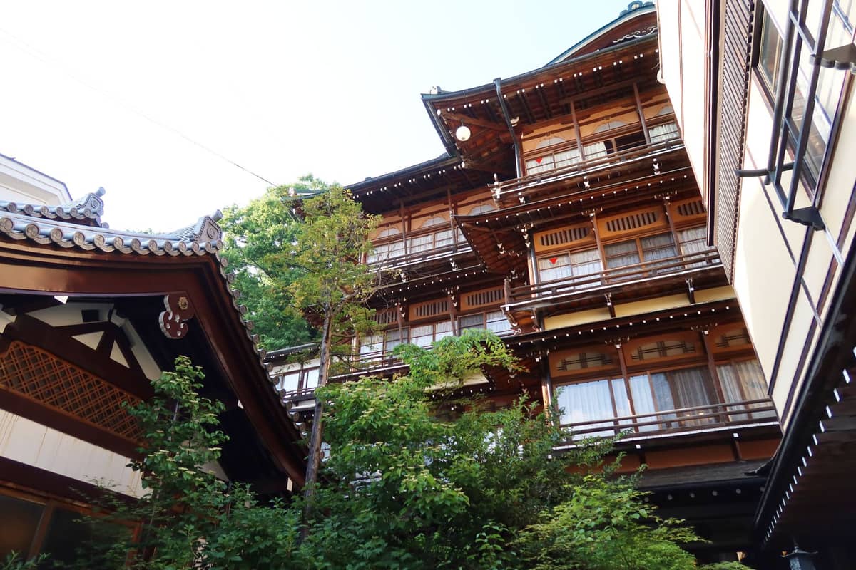 Traditional multi-story wooden Japanese bathhouse building with trees and greenery in an urban environment, viewed from a low angle.