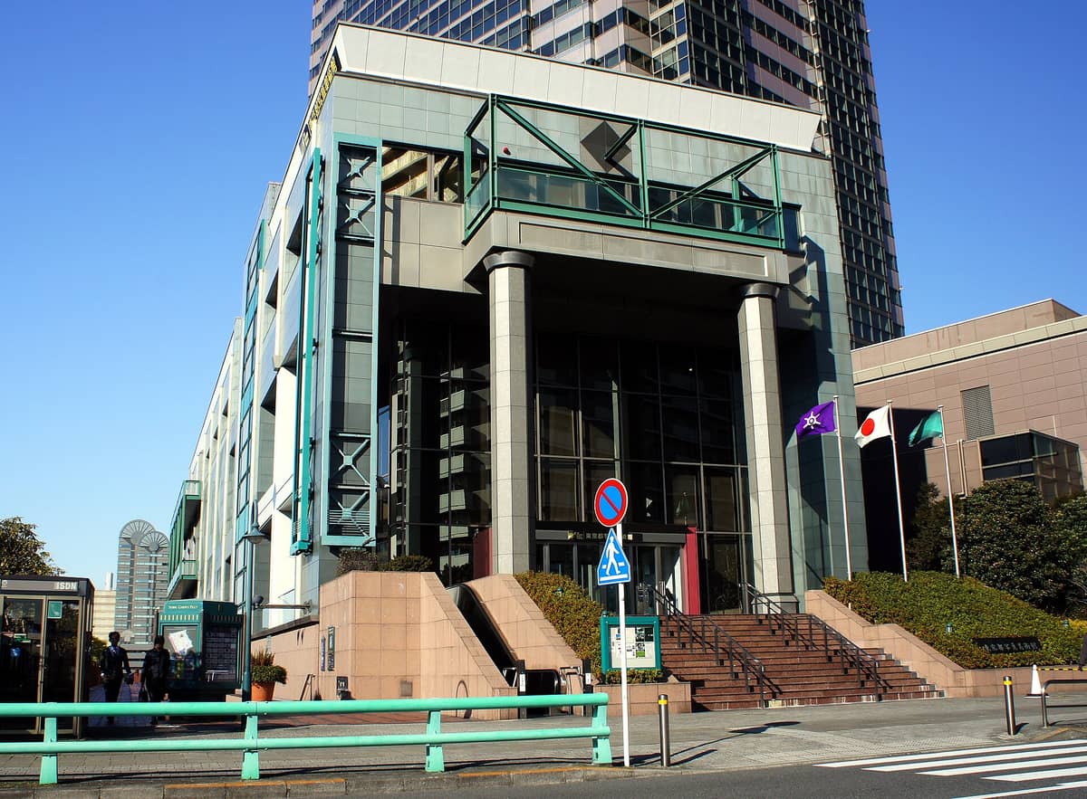 Exterior frontal view of the Tokyo Photographic Art Museum building with large columns, glass windows, and flags, located in a modern urban area.