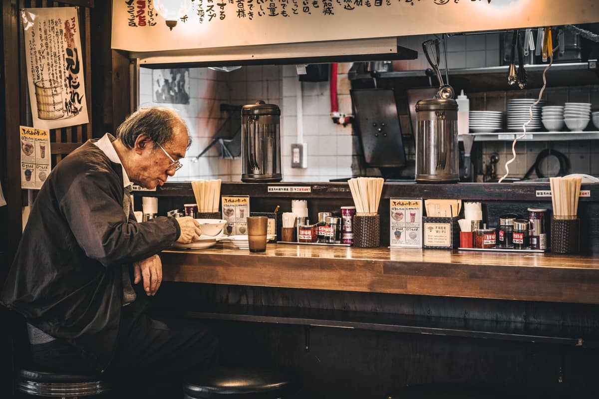 Older man eating ramen alone at a counter in a traditional Japanese ramen shop, with condiments and utensils on the counter.