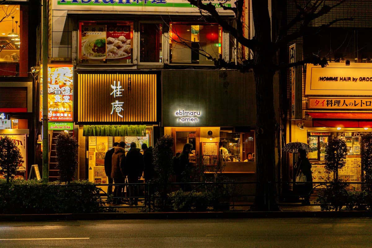 Tokyo street at night with people lined up outside a brightly lit ramen shop, storefront signs glowing in wintery darkness.