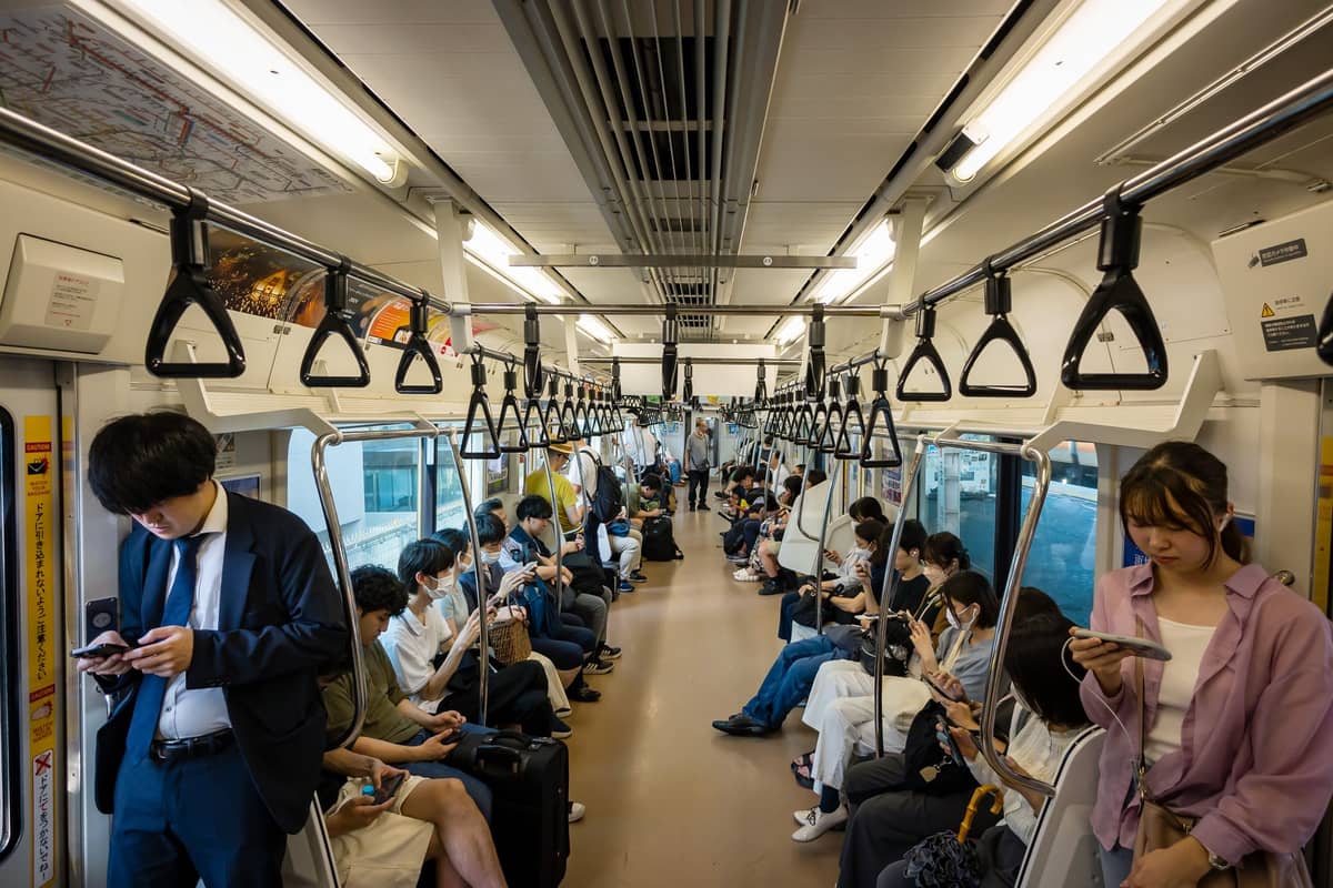 Interior of a Tokyo train at night with passengers sitting and standing, some using phones, under bright lights and hand straps.