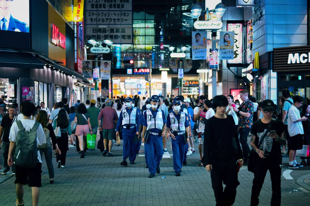 Several uniformed police officers patrol a busy pedestrian street in Tokyo at night, with many people and bright city lights in the background.