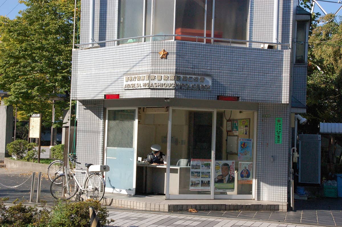 A Tokyo police box (koban) with a uniformed officer seated inside, bicycles parked outside, and signage in both Japanese and English.