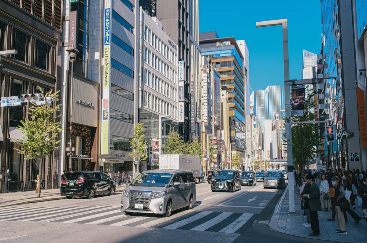 Broad daylight view of a busy Tokyo shopping street lined with modern buildings and retail stores, people walking and cars passing by.