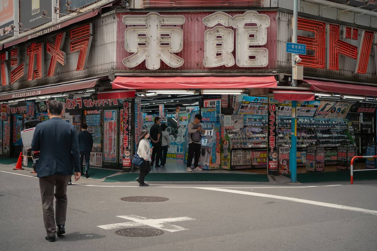 Street view of a busy Tokyo electronics store with large red and white signage, people walking and shopping outside, filled with colorful merchandise displays.