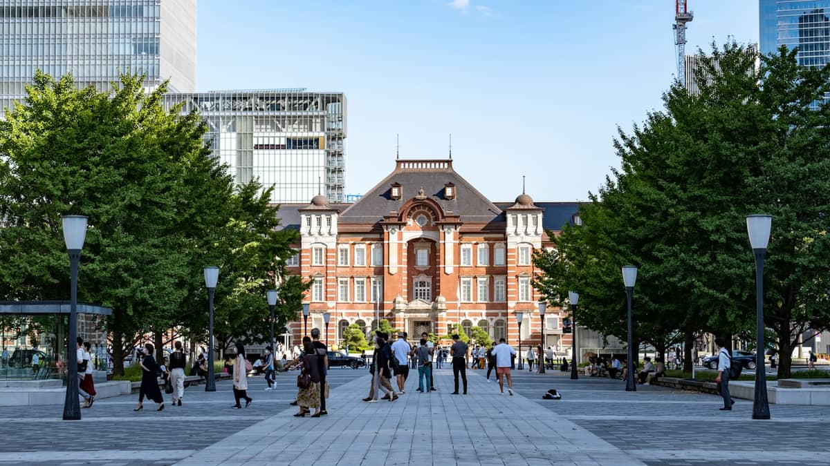 Daytime view of Tokyo Station's iconic red-brick Marunouchi facade, surrounded by people and flanked by modern skyscrapers and green trees.