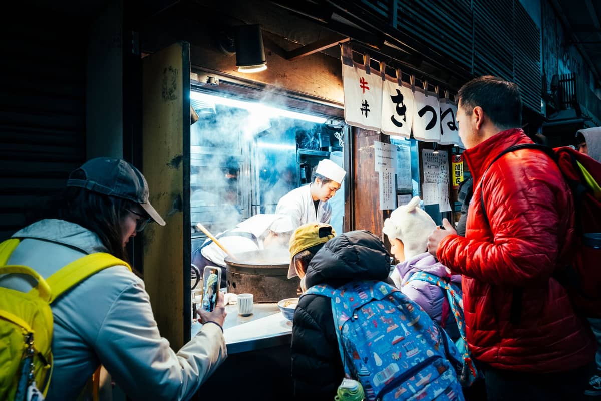 A busy Tokyo street food stall at night with a chef preparing hot food while people, including children, wait and take photos.
