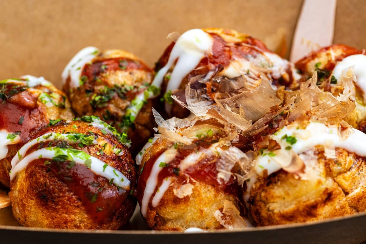 Close-up of takoyaki, Japanese octopus balls, topped with sauces and bonito flakes in a cardboard tray.