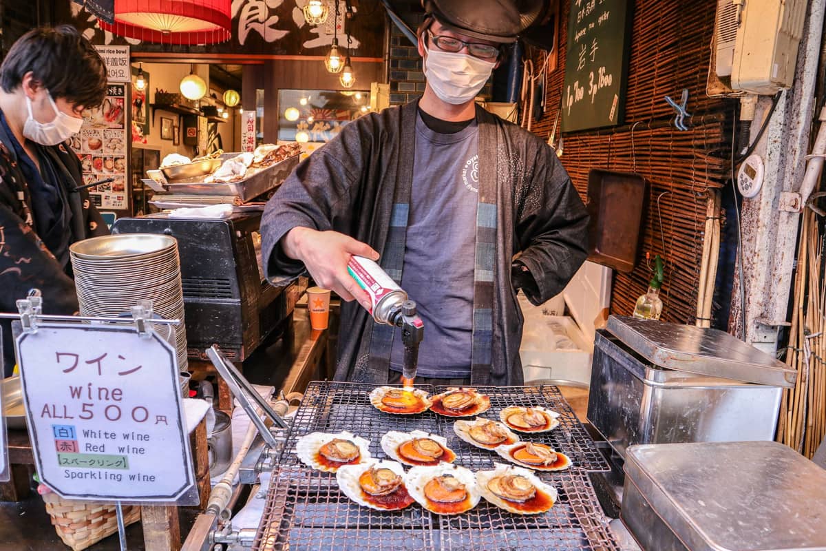 Street food vendor grilling scallops with a torch at a bustling Tokyo market, food and market stalls in background