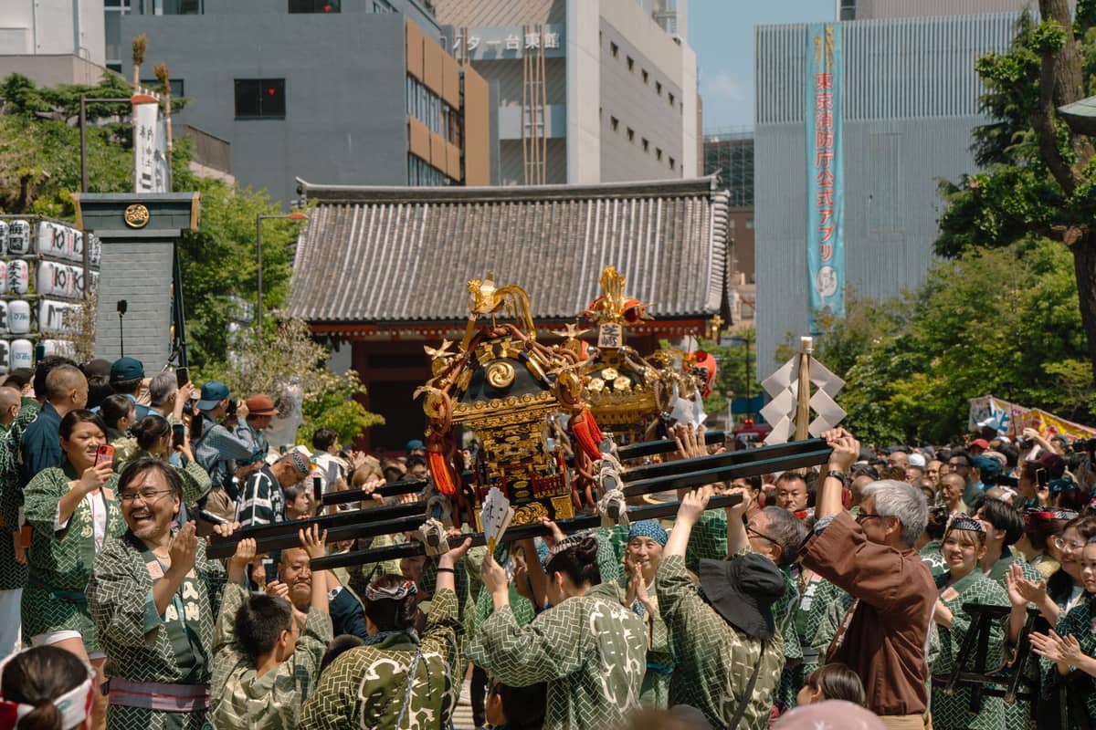 A lively summer matsuri scene in Tokyo with people carrying a mikoshi portable shrine through a city street lined with festival lanterns.