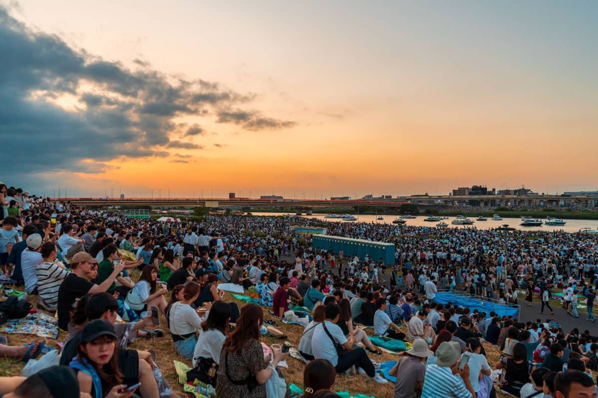 Massive crowd seated along a river at sunset, blankets spread out, waiting for a Tokyo summer festival fireworks event.