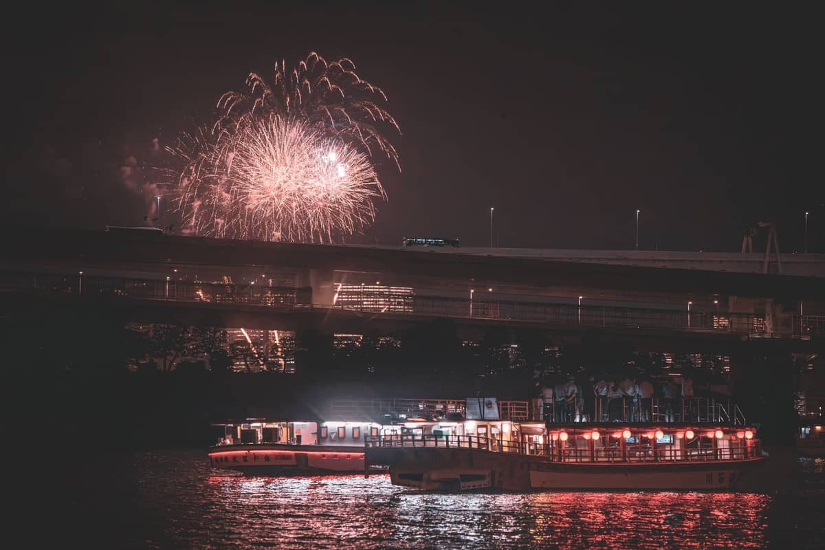 Night view of Sumida River with illuminated yakatabune boats and large fireworks bursting in the sky above a bridge in Tokyo.