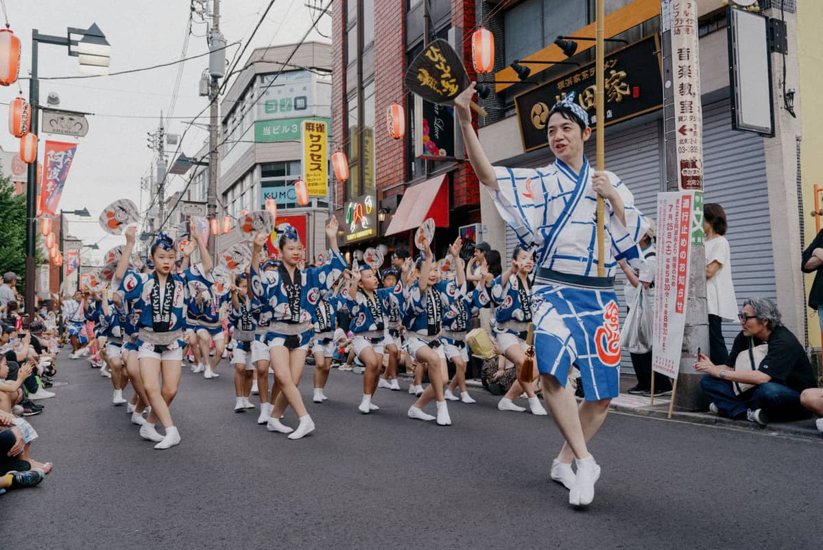 A procession of dancers in traditional costumes parades down a Tokyo street during a summer festival, with crowds lining the sidewalks.