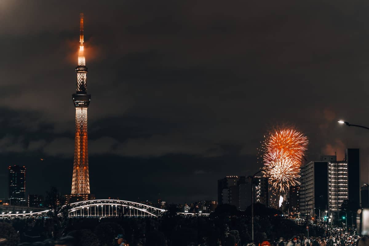 Tokyo Skytree tower at night with colorful fireworks bursting over city buildings and a large crowd gathered along the river for a summer festival.