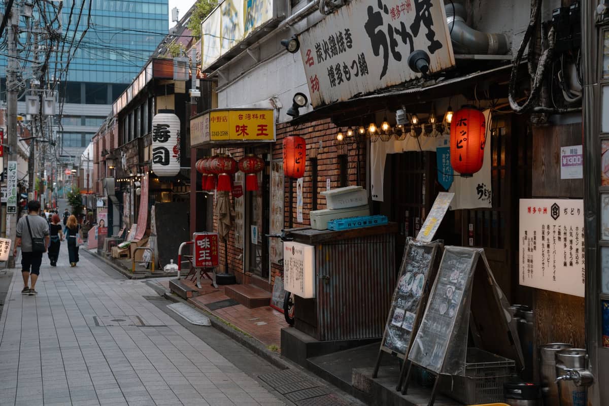Narrow street in Tokyo lined with small restaurants and izakaya, with lanterns and signs, early evening with a few pedestrians