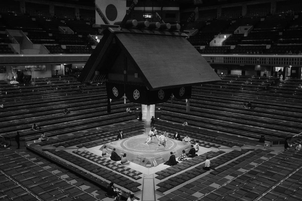 View of Ryogoku Kokugikan sumo arena with early-day low crowds and sumo wrestlers in the center ring preparing for matches.