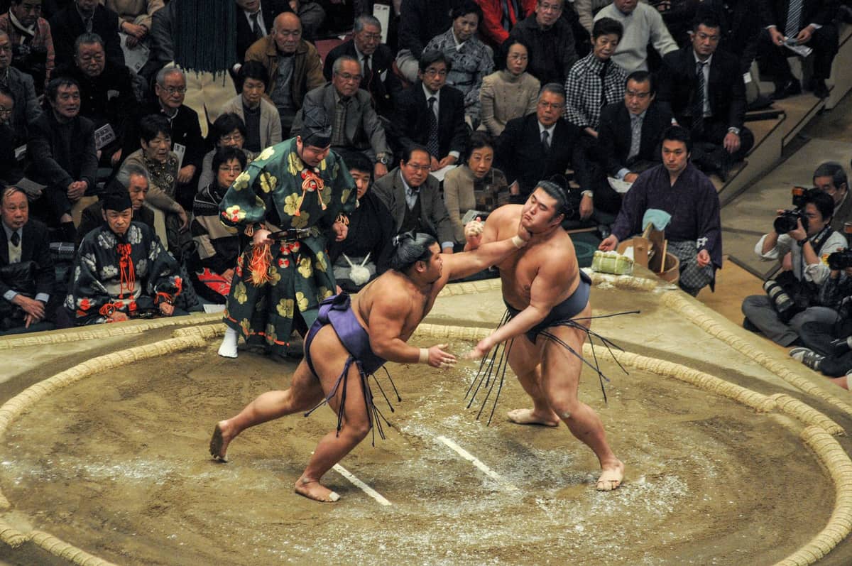 Sumo wrestling match in progress, with two wrestlers in the ring surrounded by a large crowd of spectators at an indoor arena.