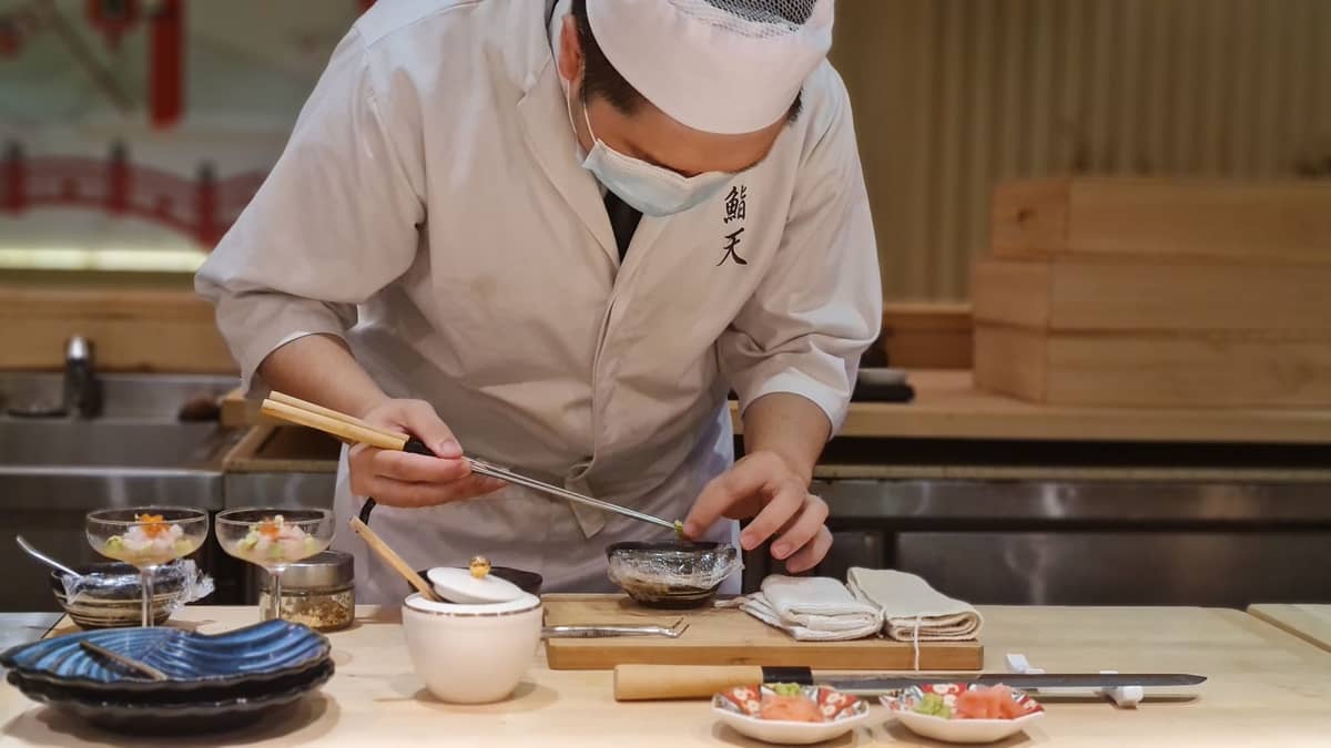 Japanese chef in traditional uniform prepares dishes at a sushi counter, with small bowls and plates arranged in front of him.