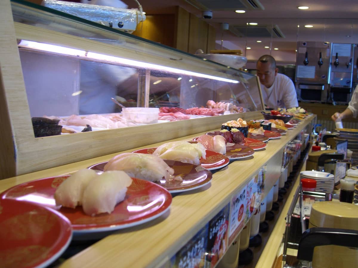 A sushi chef behind a counter at a conveyor belt sushi restaurant in Japan, with many colorful sushi plates moving past customers.