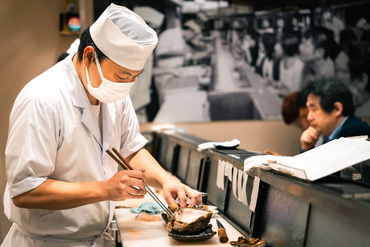 A sushi chef in traditional attire prepares sushi at a counter with diners in the background, inside a Tokyo sushi restaurant.