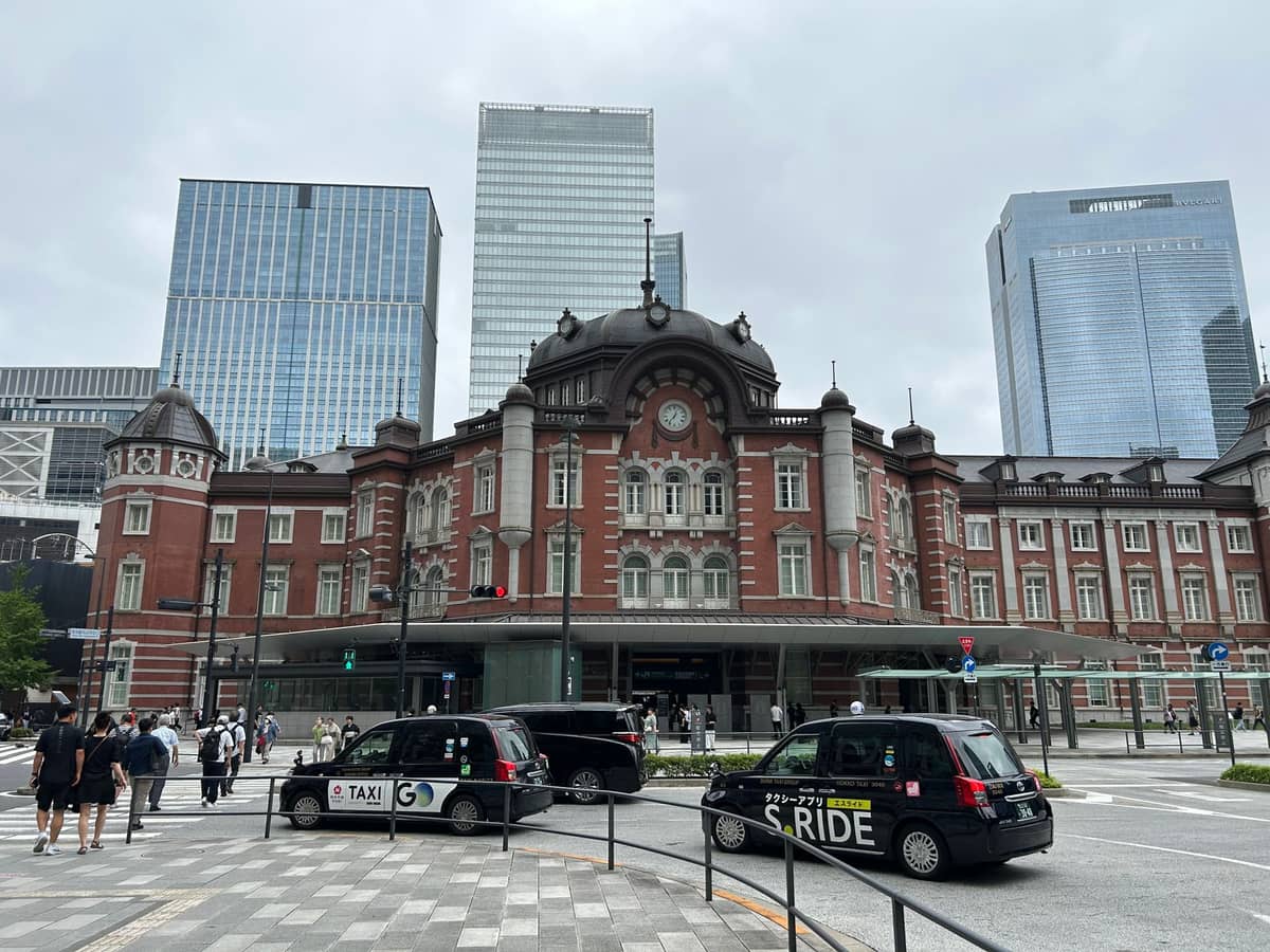 Daytime view of Tokyo Station’s iconic red-brick Marunouchi building with city skyscrapers in the background and taxis in the foreground.