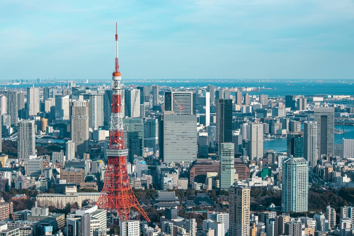 A clear, wide daytime view of Tokyo Tower rising above the surrounding city skyline, with buildings and Tokyo Bay in the background.