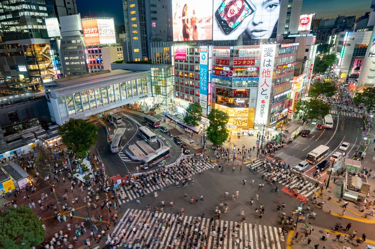 Aerial view of the busy Shibuya Crossing at dusk, surrounded by illuminated buildings, crowds of pedestrians, and city lights in central Tokyo.