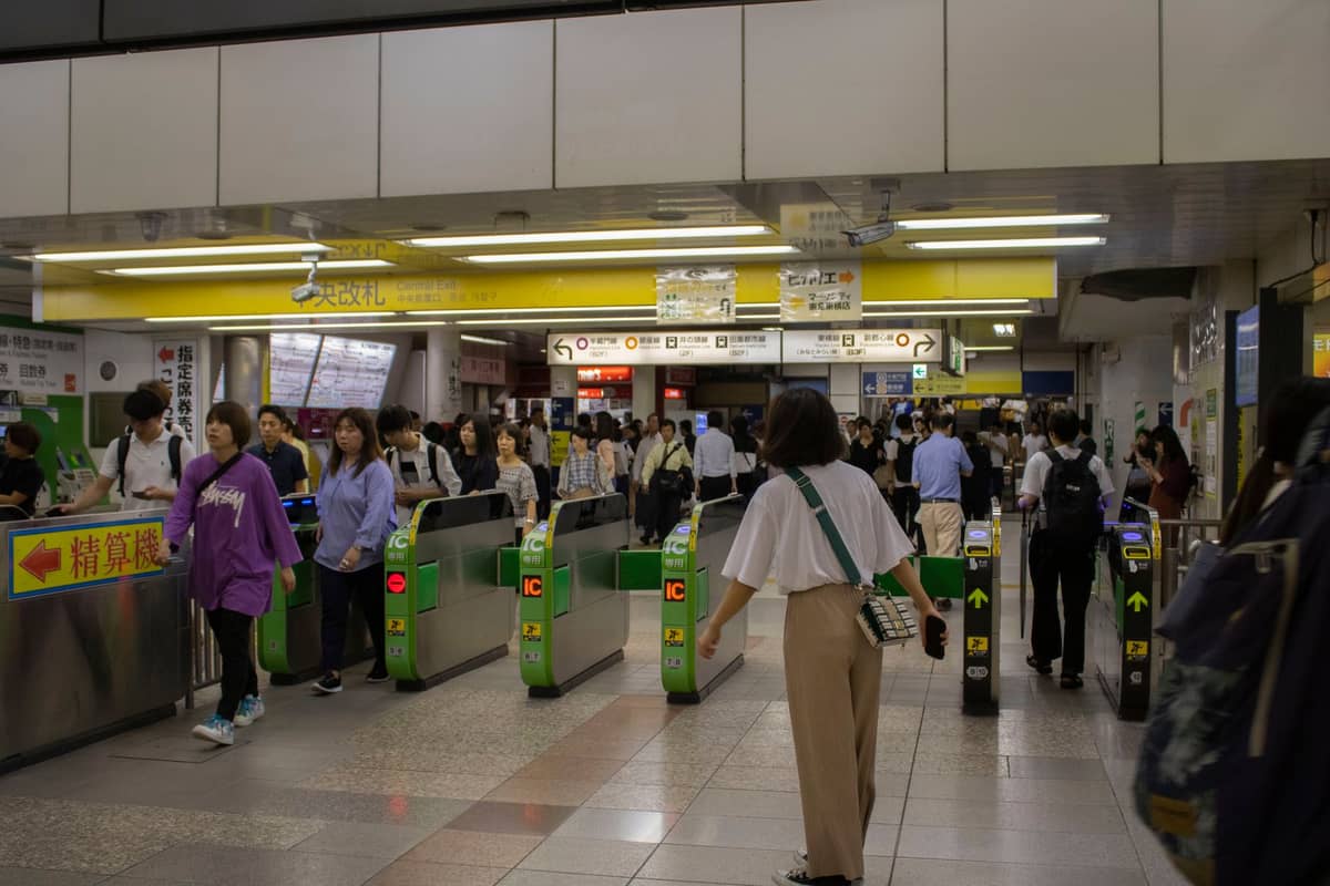 Crowded Tokyo train station entrance with green ticket gates, people using IC cards, and Japanese signage above the barriers.
