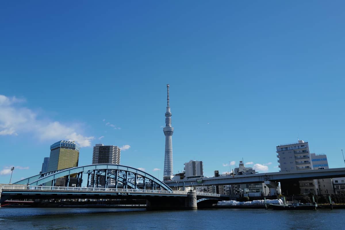 Tokyo Skytree rising above the Sumida River, with pedestrian bridges and waterfront promenades under a clear blue sky.