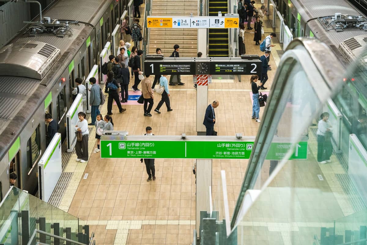 A busy Tokyo train station interior with Yamanote Line trains, green signage, and people navigating platforms and escalators.