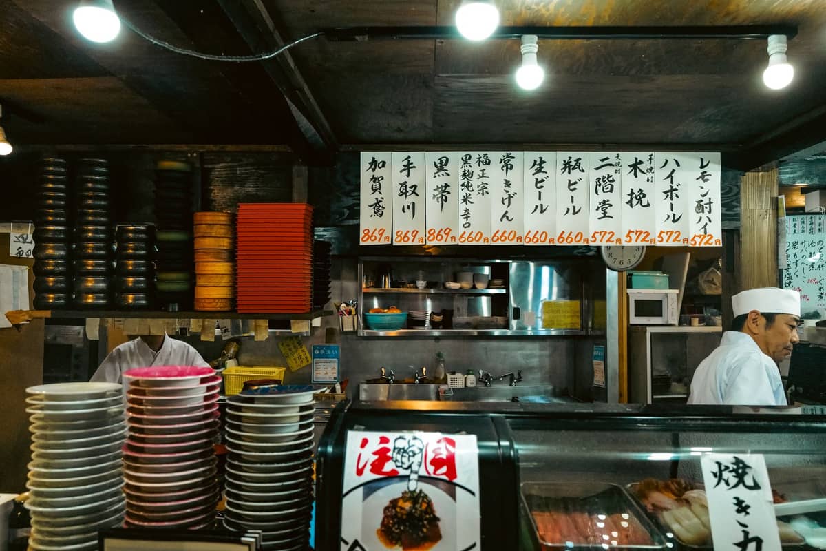 Interior of a Japanese sushi restaurant in Tokyo with chefs, counter seating, and handwritten menu signs above the kitchen area.