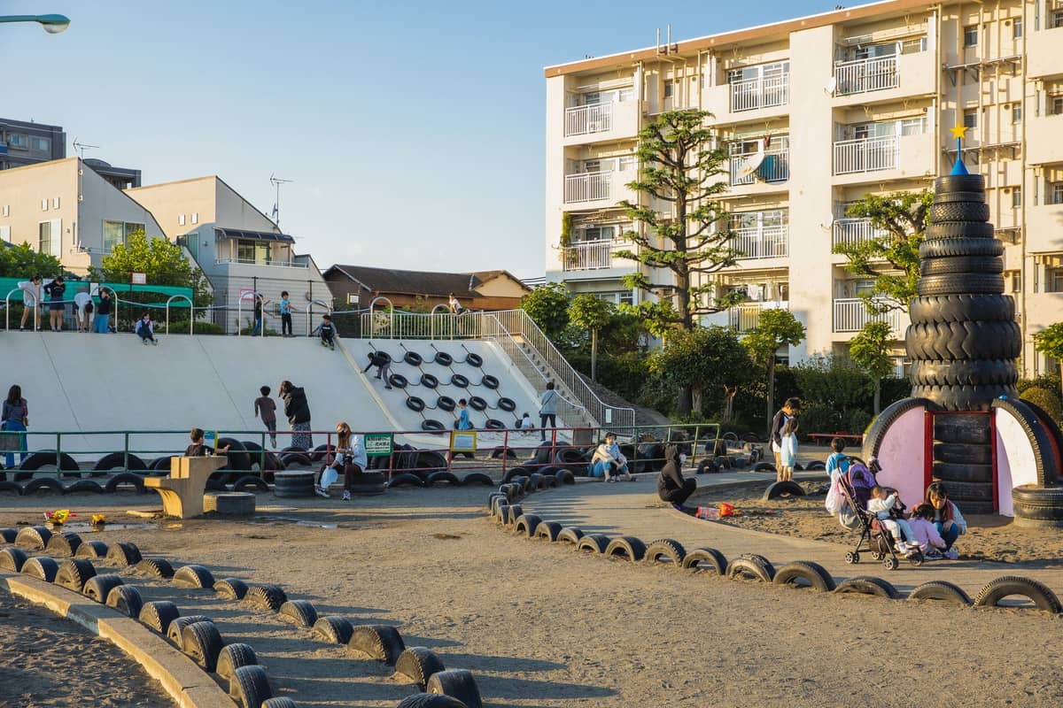 Large playground in Tokyo with families and children playing, featuring a unique tire structure and apartment buildings in the background.