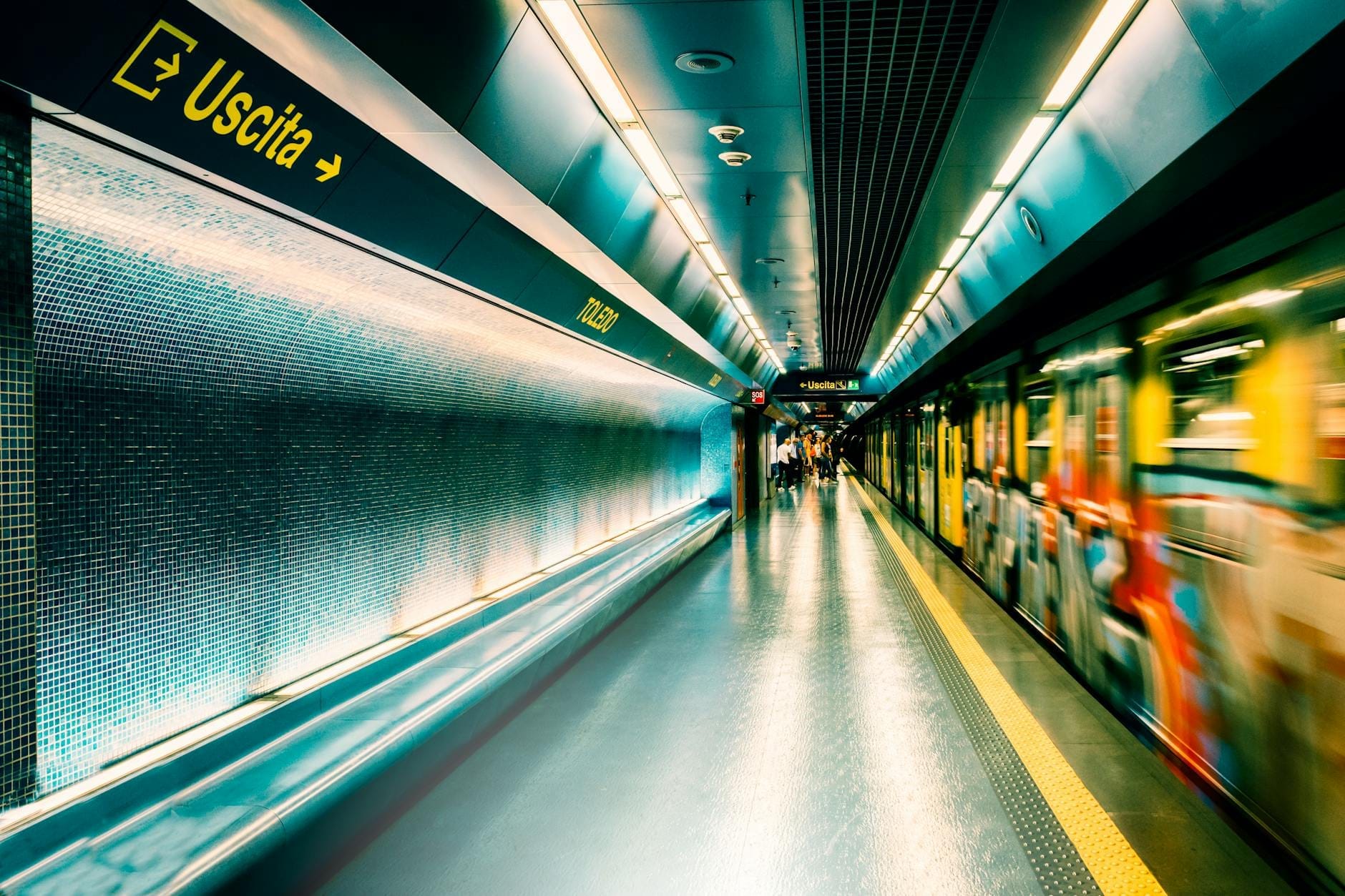 Banchina di una moderna stazione della metropolitana con illuminazione dinamica, parete a mosaico blu, cartello con la scritta 'Uscita' e un treno in movimento che crea un'atmosfera artistica sotterranea.
