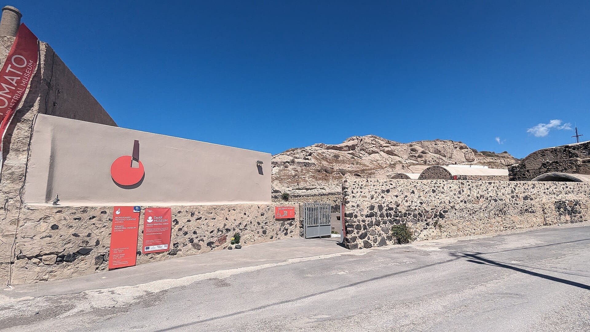 ทางเข้าหลักของ Tomato Industrial Museum D. Nomikos ใน Santorini พร้อมกำแพงหิน ป้ายสีแดง และเนินเขาหินภายใต้ท้องฟ้าสีฟ้าใส