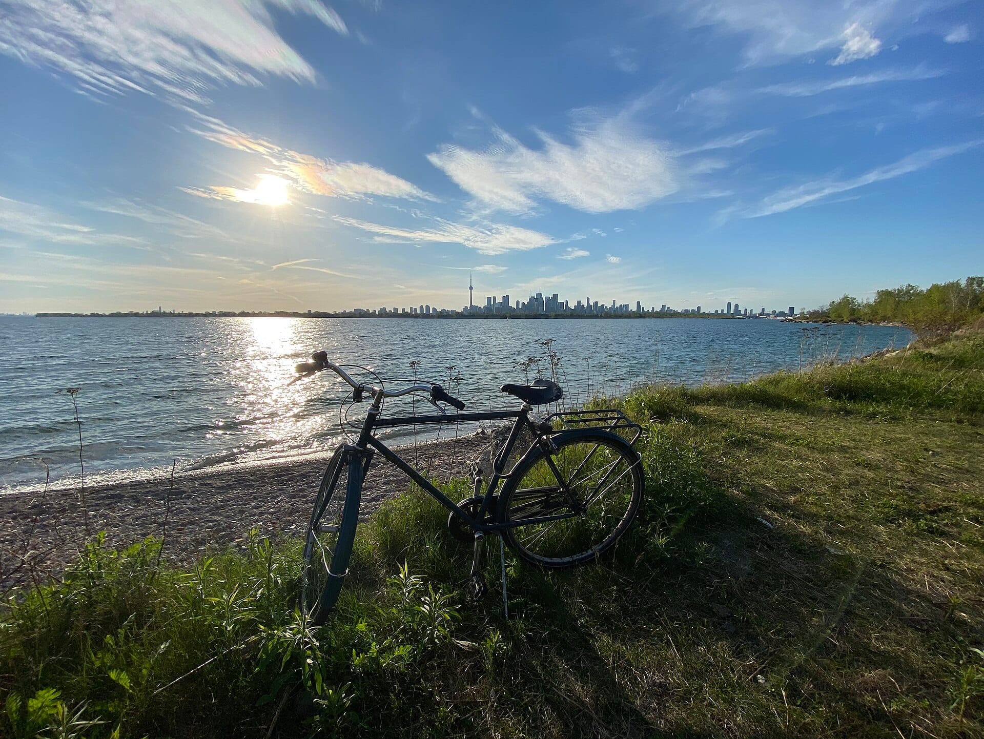 จักรยานพิงอยู่บนชายฝั่งที่เต็มไปด้วยหญ้าของ Tommy Thompson Park พร้อมเส้นขอบฟ้าของโตรอนโตและทะเลสาบออนแทรีโอภายใต้ท้องฟ้าที่มีเมฆบางส่วนและแสงแดด