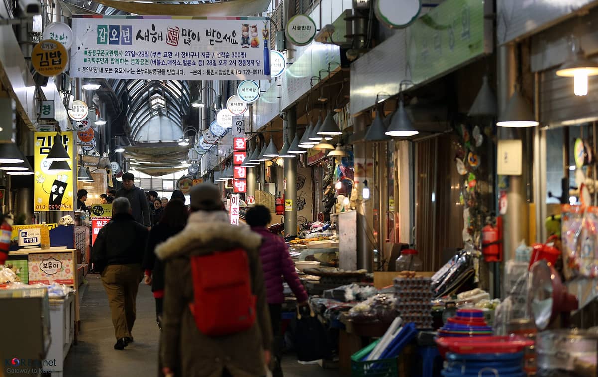 O movimentado corredor interno do Mercado Tongin em Seul, com bancas de comida, compradores e faixas coloridas penduradas sob as luzes do mercado.