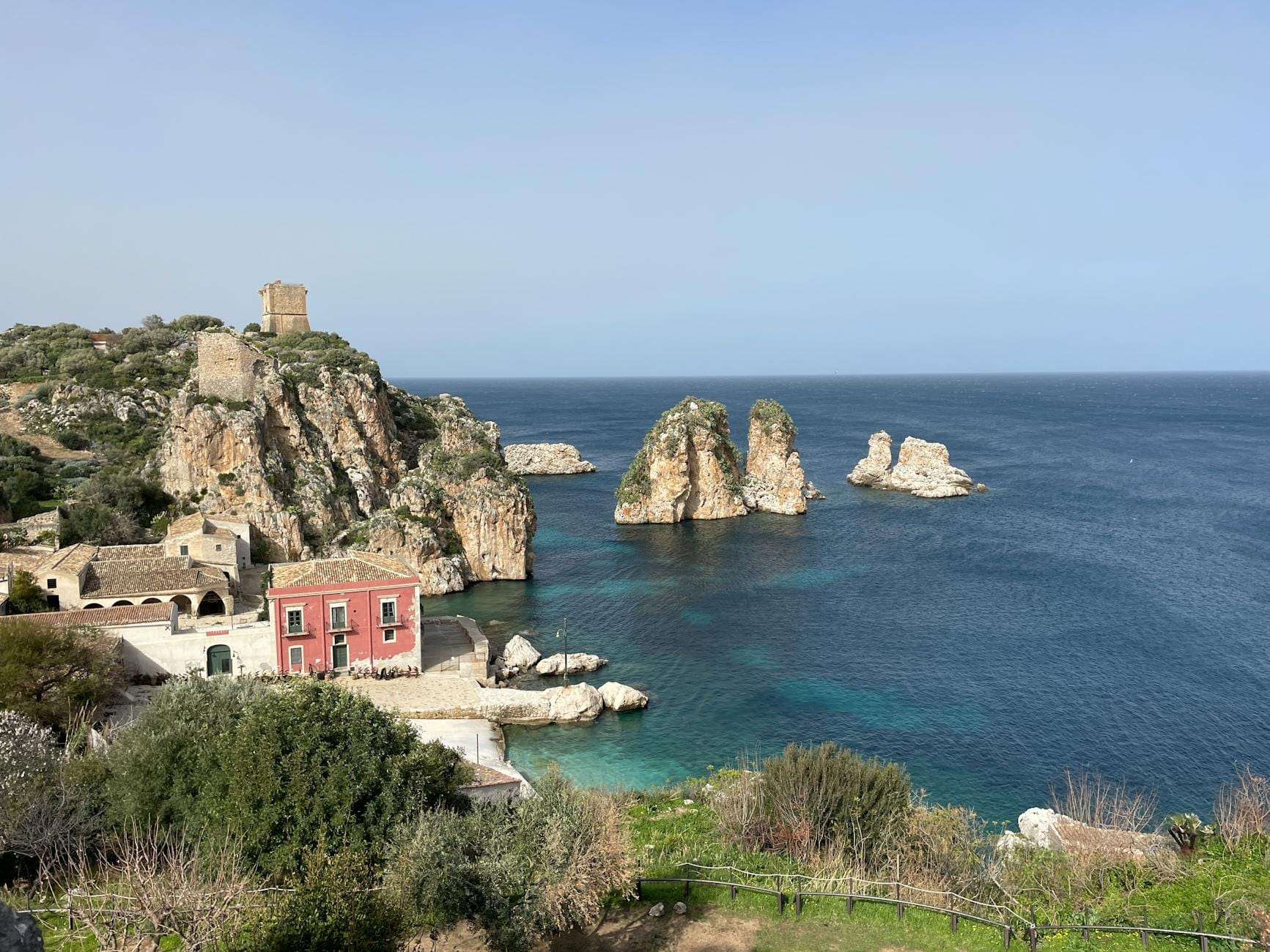 Malerischer Blick auf die Tonnara di Scopello mit historischen Steingebäuden, aus dem klaren blauen Wasser ragenden Felsnadeln und einer dramatischen Küstenlinie im hellen Tageslicht.