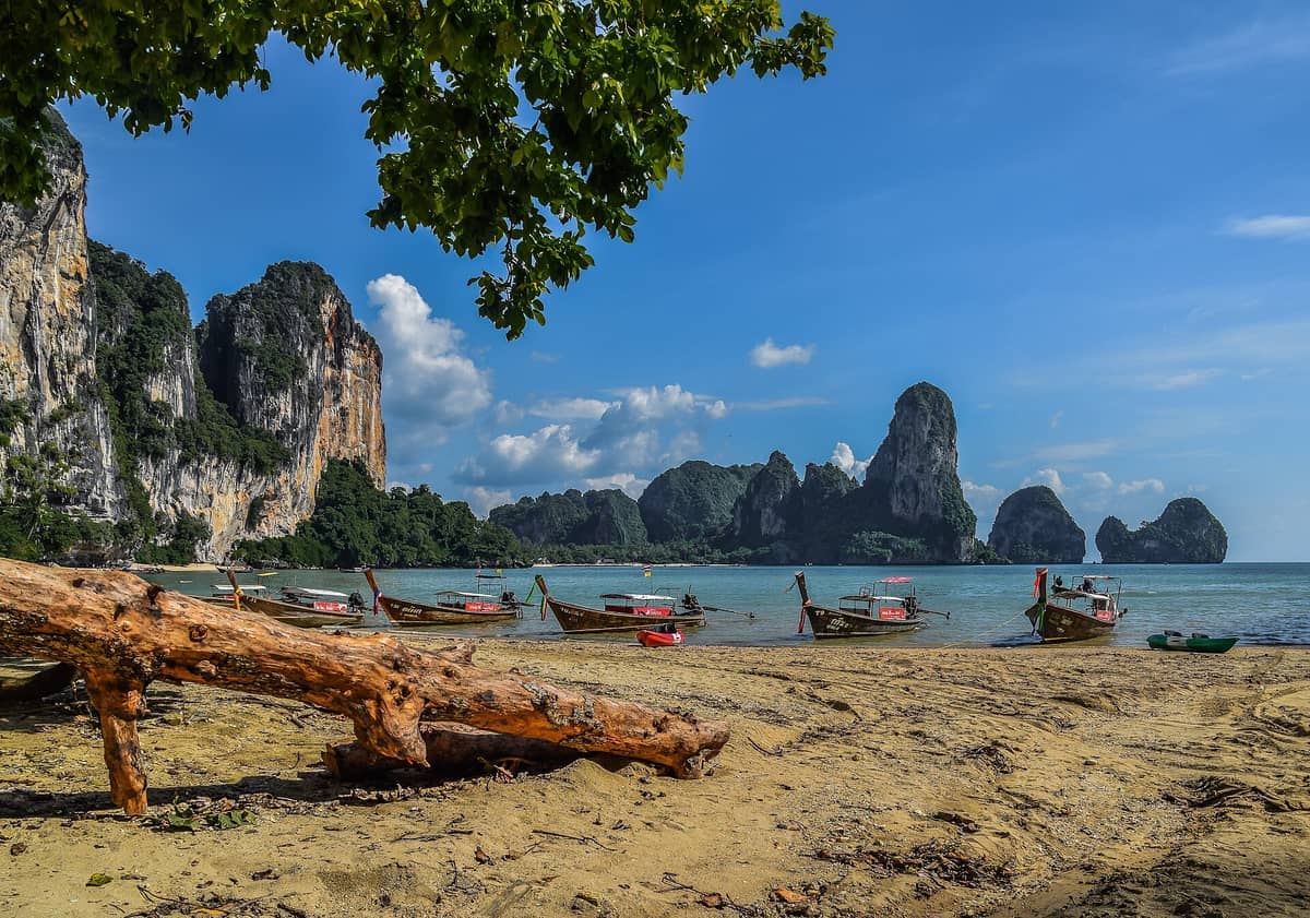 Tonsai Beach in Krabi with longtail boats anchored along sandy shore, dramatic limestone cliffs, clear blue sky, and lush green foliage visible.