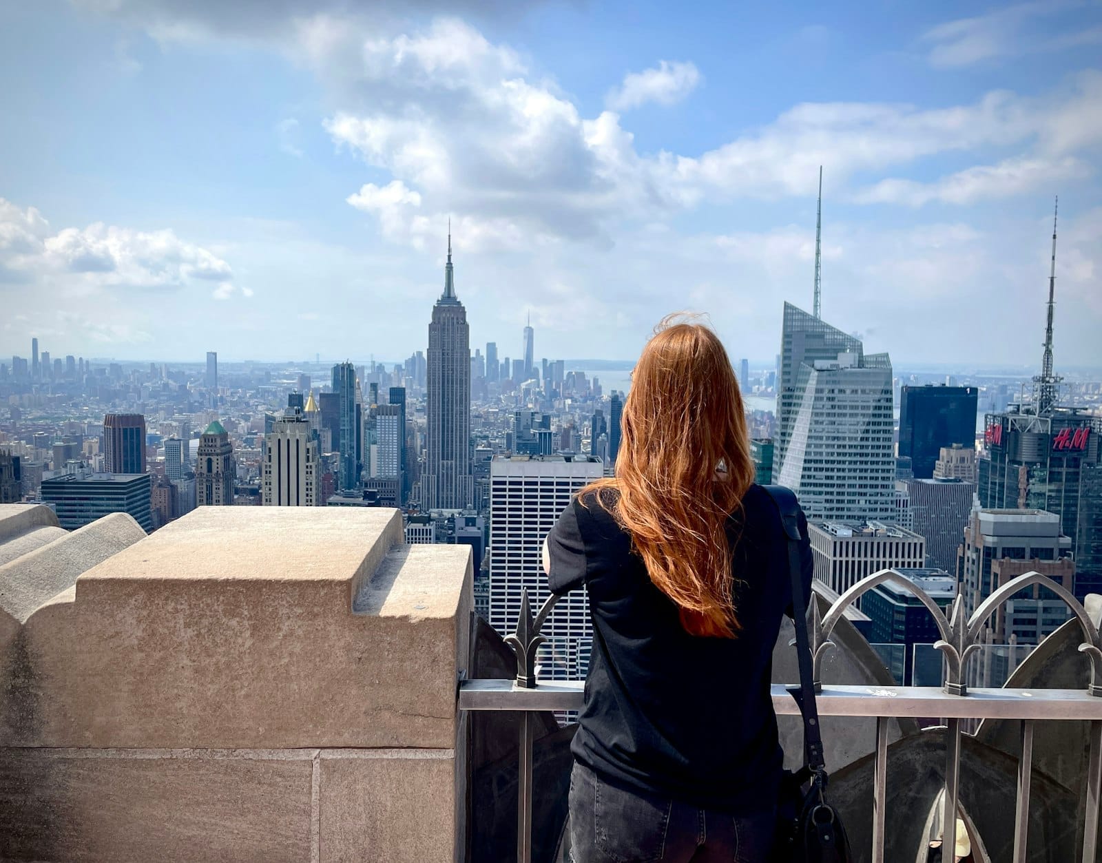 Una donna con i capelli rossi osserva lo skyline di Manhattan dal Top of the Rock, con l'Empire State Building ben visibile in una giornata parzialmente nuvolosa.