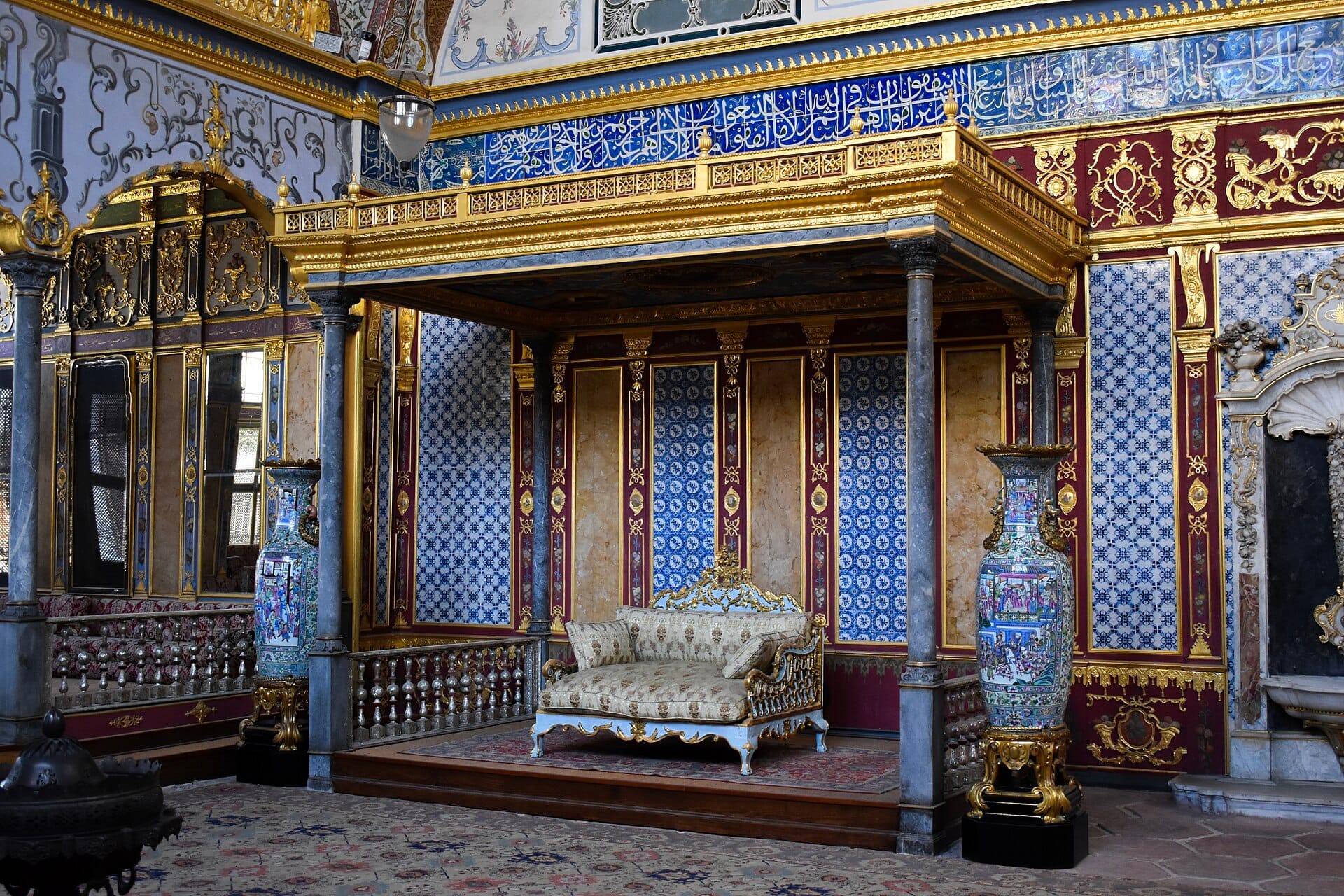 La sontuosa sala delle udienze nell'Harem del Palazzo Topkapı con decorazioni dorate, piastrelle blu a motivi, colonne di marmo e un elegante divano centrale.