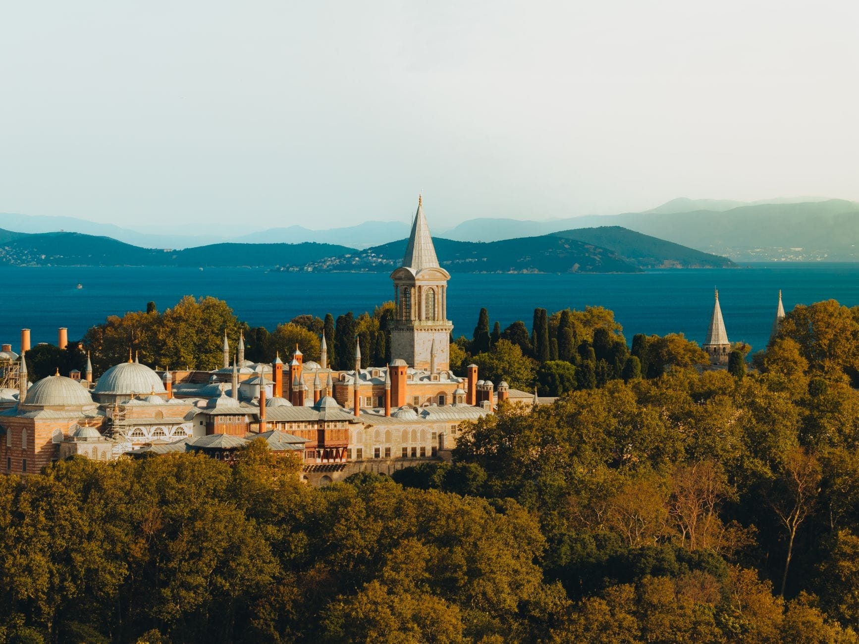 Vista panoramica del Palazzo di Topkapı con cupole e torre che si innalzano sopra una rigogliosa vegetazione, sullo sfondo delle acque blu scintillanti e delle colline lontane di Istanbul.