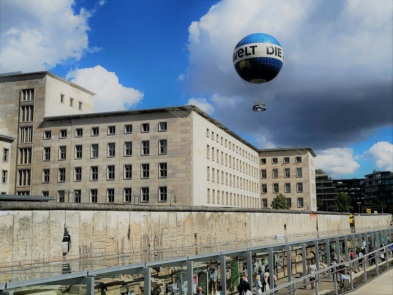 Edificio storico in pietra e tratto conservato del Muro di Berlino presso il sito della Topography of Terror, con una mongolfiera che galleggia sotto un cielo parzialmente nuvoloso.