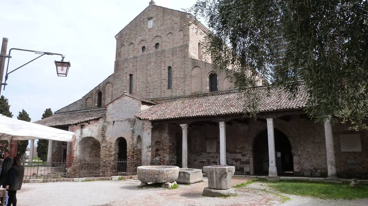 วิวภายนอกของ Basilica di Santa Maria Assunta บนเกาะ Torcello พร้อมด้วยผนังอิฐโบราณ หน้าต่างโค้งโคง เสาหิน และลักษณะลานภายนอกที่เก่าแก่