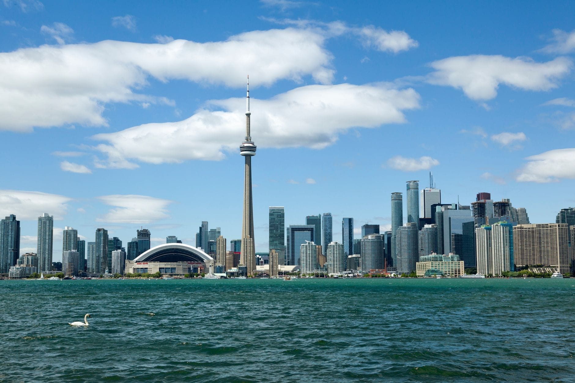 Skyline de Toronto com a CN Tower em um dia ensolarado, vista de além do lago com céu azul e nuvens brancas.