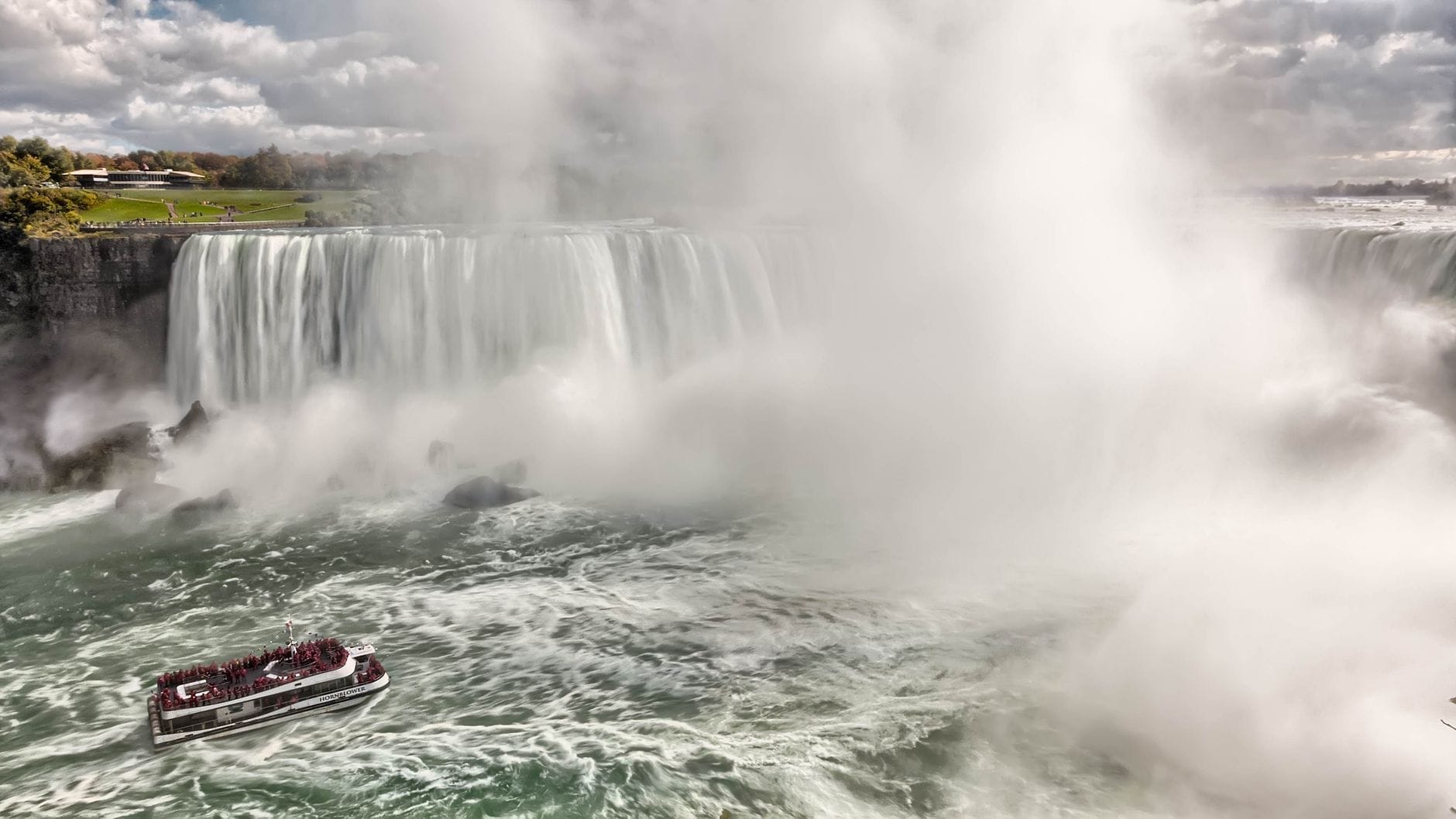 Vista ampla das Cataratas do Niágara com um barco turístico em primeiro plano e névoa subindo das poderosas quedas d'água.