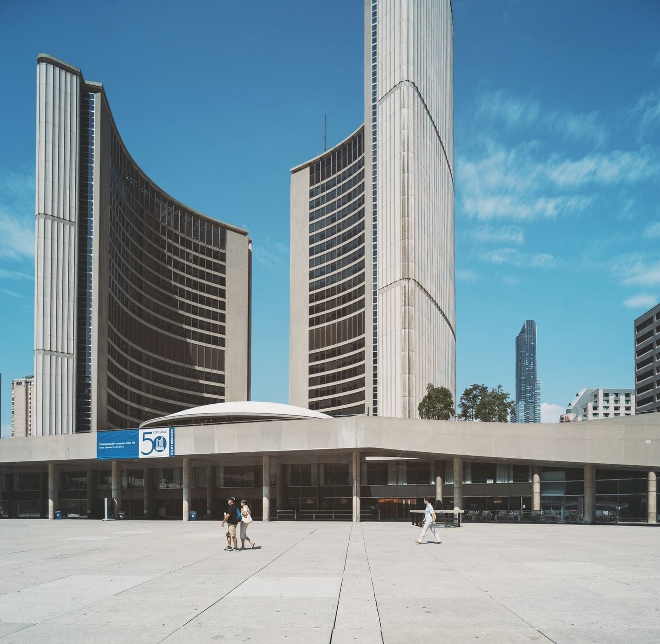 A Câmara Municipal de Toronto num dia ensolarado com suas distintas torres curvas e pessoas caminhando pela Nathan Phillips Square.